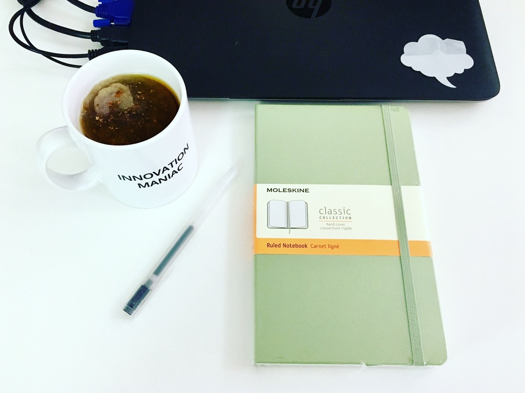 Innovation Maniac mug and brand new sage green moleskin notebook in front o fa black PC laptop with a cloud post it on the corner and a pen on a white desk.
